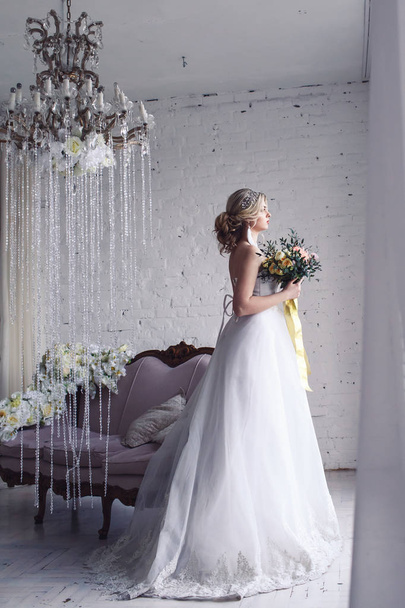 beautiful bride in white wedding dress is standing by window and holding bouquet. Morning of bride - Foto, Imagen