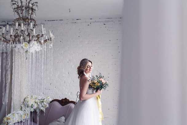 beautiful bride in white wedding dress is standing by window and holding bouquet. Morning of bride - Foto, Imagen