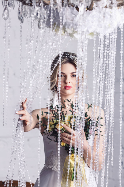 bella ragazza in abito da sposa è in piedi vicino a tende di cristallo e tenendo bouquet
 - Foto, immagini