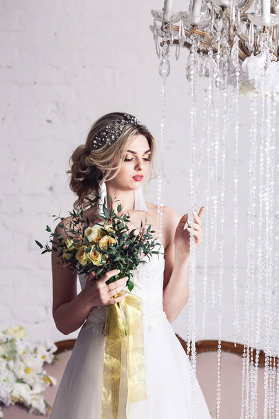 krásná dívka ve svatebních šatech stojící poblíž křišťálové závěsy a držení kytice - Fotografie, Obrázek