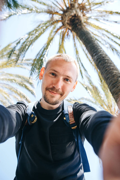 Un joven turista o bloguero hace una selfie o informa a sus suscriptores sobre el fondo de las palmeras en un país caliente
 - Foto, imagen