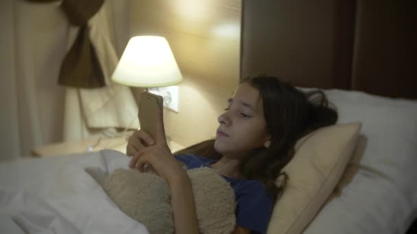 Cute little girl in her room at night, lying on bed using smartphone. her night lamp is on. 4k. - Кадры, видео
