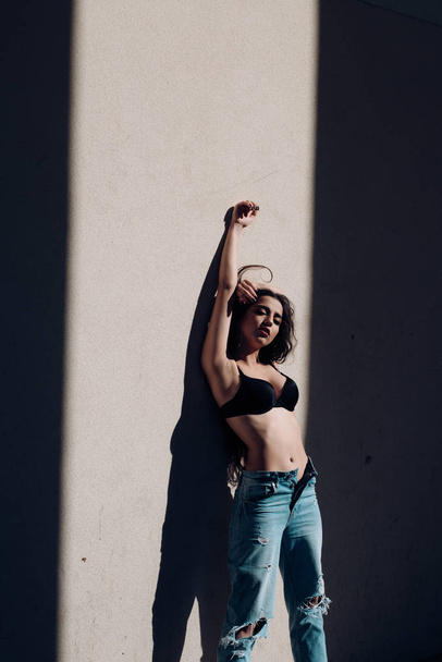 Slim beautiful girl in black bra and blue denim jeans on white background. Sexy brunette leaning on wall outdoors, passion and seduction concept. - Foto, afbeelding
