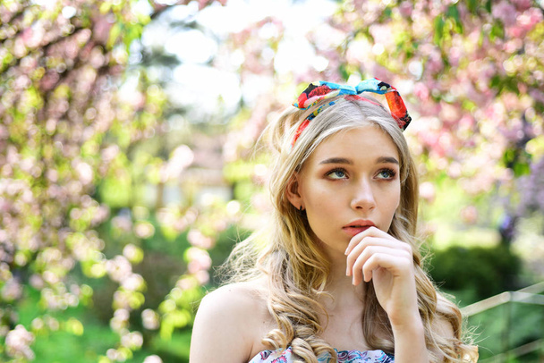 Thoughtful girl in retro outfit with colorful headband dreaming in floral garden, unity with nature concept. Lovely young lady strolling between blooming trees on sunny day. - Zdjęcie, obraz