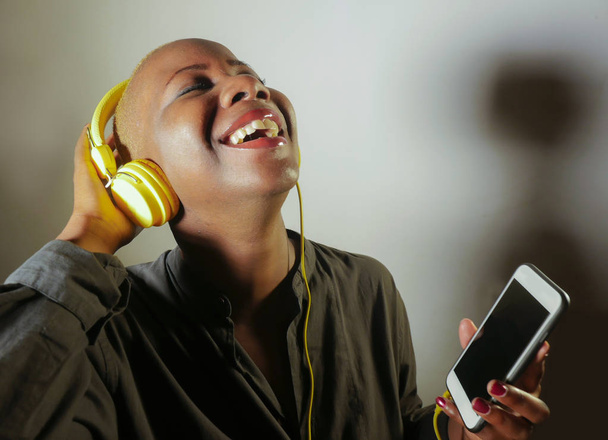 young happy and attractive afro American woman with yellow headphones and mobile phone listening to internet music song smiling cheerful isolated on grey green background in hipster lifestyle - Fotó, kép