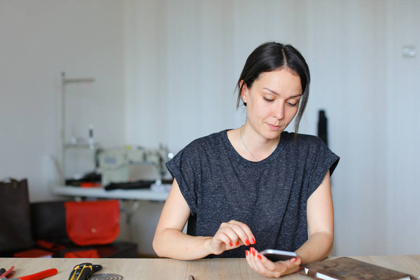 Ευρωπαϊκή craftswoman κάθεται στο ατελιέ και χρησιμοποιώντας το smartphone, χειροποίητα δερμάτινα είδη. - Φωτογραφία, εικόνα