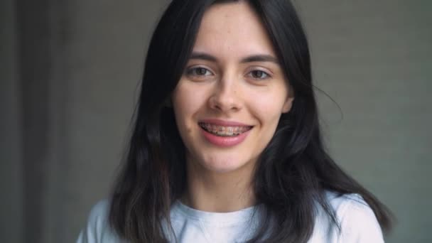 A young girl with braces on her teeth tilts her head and appears in the frame. Young woman on white wall background smiling with braces - Πλάνα, βίντεο