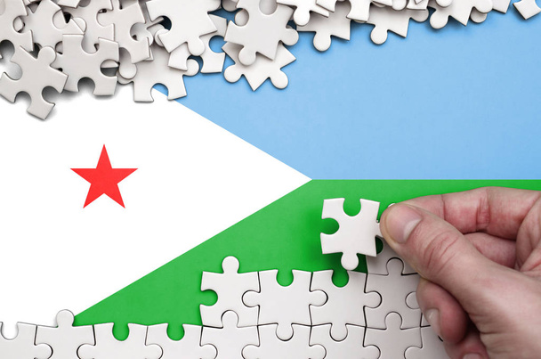 Dschibuti-Flagge ist auf einem Tisch abgebildet, auf dem die menschliche Hand ein Puzzle weißer Farbe faltet. - Foto, Bild