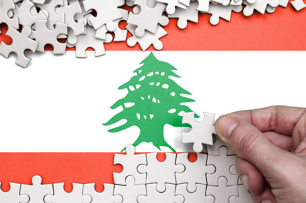 Σημαία του Λιβάνου απεικονίζεται σε έναν πίνακα στον οποίο το ανθρώπινο χέρι διπλώνει ένα παζλ από λευκό χρώμα. - Φωτογραφία, εικόνα
