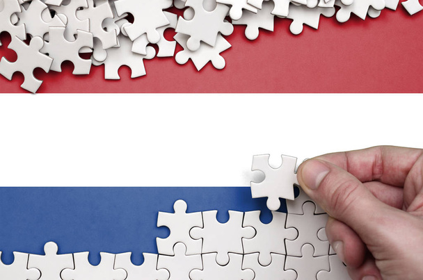 Bandera de los Países Bajos se representa en una mesa en la que la mano humana pliega un rompecabezas de color blanco
. - Foto, Imagen