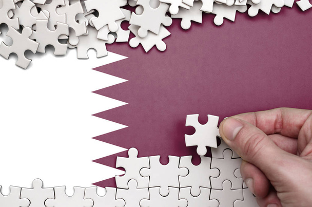 Σημαία του Κατάρ απεικονίζεται σε έναν πίνακα στον οποίο το ανθρώπινο χέρι διπλώνει ένα παζλ από λευκό χρώμα. - Φωτογραφία, εικόνα