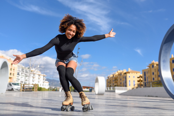 Young passen zwarte vrouw op rolschaatsen riding buitenshuis op stedelijke straat met open armen. Lachende meisje met afro kapsel skaten op zonnige dag - Foto, afbeelding