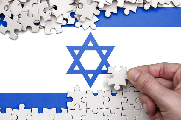 Bandera de Israel se representa en una mesa en la que la mano humana pliega un rompecabezas de color blanco
. - Foto, Imagen