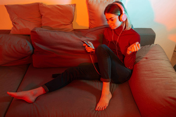 Pretty lady sitting on sofa with headphones and cellphone in hands. Young woman listening music in headphones while spending time at home - 写真・画像