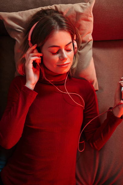 Портрет леді в навушниках лежить на дивані з мобільним телефоном в руці і слухає музику вдома
 - Фото, зображення
