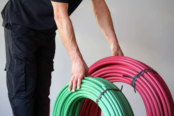 The electrician holds green and red a roll of an electric protective tube on a gray background. The concept of industrial and private services. - Photo, Image