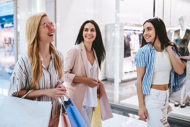 Drie aantrekkelijke jonge meisjes doen winkelen met boodschappentassen in moderne winkelcentrum. - Foto, afbeelding
