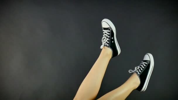 Ženské nohy v klasické černé a bílé tenisky. Nohy jsou vyvolány nahoru a houpat ze strany na stranu. Šedé, černé pozadí. Klasické boty, retro styl. Zdravý životní styl - Záběry, video