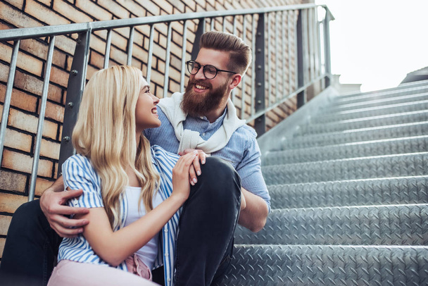 Young stylish happy couple is spending time together outdoors. Attractive woman and handsome bearded man are sitting on stairs in the city. - Φωτογραφία, εικόνα