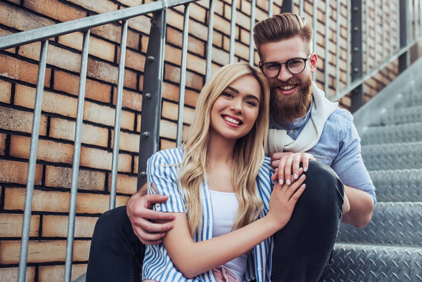 Young stylish happy couple is spending time together outdoors. Attractive woman and handsome bearded man are sitting on stairs in the city. - Fotó, kép