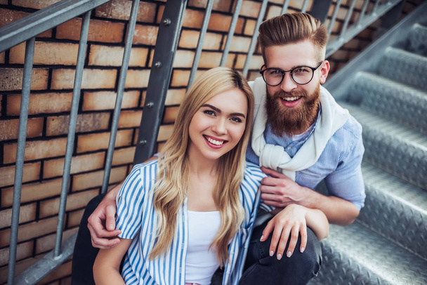 Young stylish happy couple is spending time together outdoors. Attractive woman and handsome bearded man are sitting on stairs in the city. - Фото, зображення