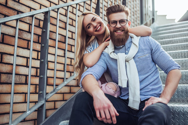 Young stylish happy couple is spending time together outdoors. Attractive woman and handsome bearded man are sitting on stairs in the city. - Foto, Imagem