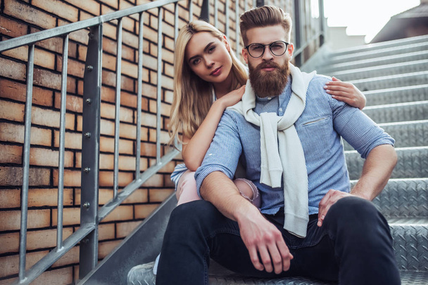 Young stylish happy couple is spending time together outdoors. Attractive woman and handsome bearded man are sitting on stairs in the city. - Foto, afbeelding