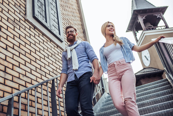 Young stylish happy couple is spending time together outdoors. Attractive woman and handsome bearded man are walking on stairs in the city. - Valokuva, kuva