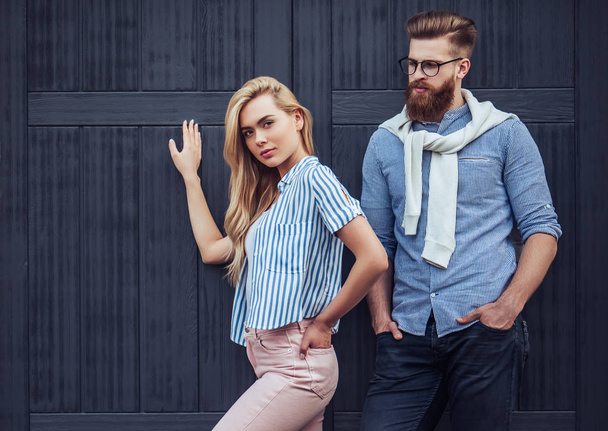 Stylish young couple is spending time together outdoors. Attractive woman and handsome bearded man are posing on black wooden background. - Фото, зображення