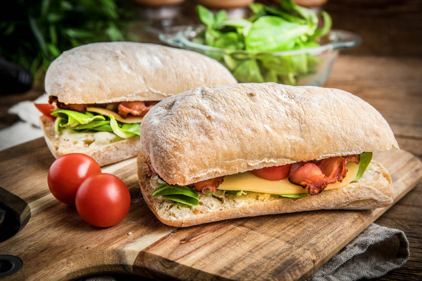 Ciabatta sandwich with arugula salad, bacon and yellow cheese on wooden board. - Photo, image