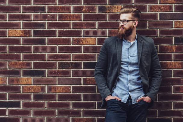 Stylish bearded man is posing on brick background in the city. Young man in eye glasses isolated. - Foto, afbeelding