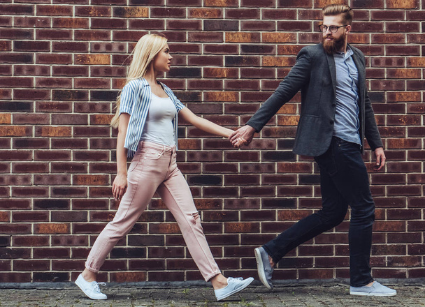 Stylish young couple is spending time together outdoors. Attractive woman and handsome bearded man are walking on brick background and holding hands. - Foto, Imagem