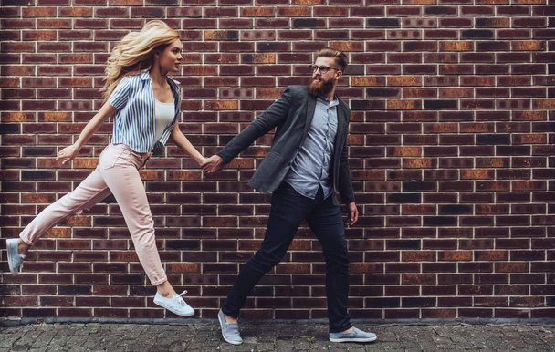 Stylish young couple is spending time together outdoors. Attractive woman and handsome bearded man are walking on brick background and holding hands. - Φωτογραφία, εικόνα