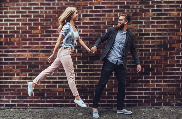 Stylish young couple is spending time together outdoors. Attractive woman and handsome bearded man are walking on brick background and holding hands. - Fotografie, Obrázek