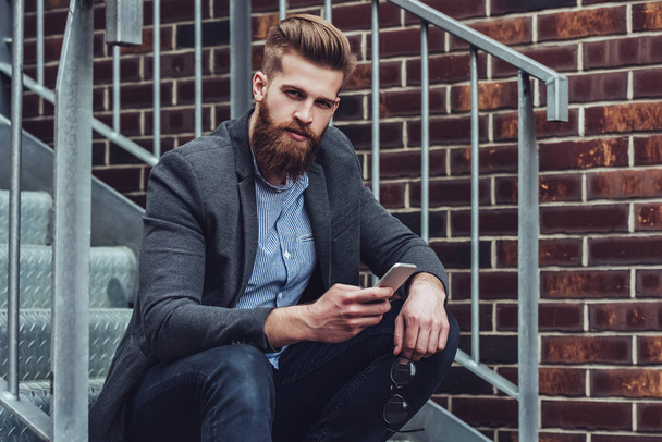 Handsome bearded man is sitting on stairs with smart phone in hands outdoors. Stylish man in the city. - Fotó, kép