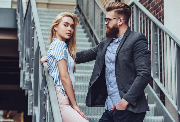 Attractive young happy couple is spending time together outdoors. Attractive woman and handsome bearded man are standing on stairs in the city. - Photo, Image