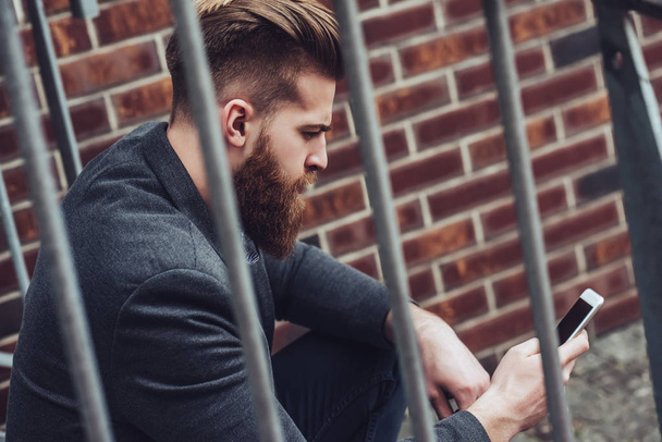 Красивый бородатый мужчина сидит на лестнице со смартфоном в руках на улице. Стильный человек в городе
. - Фото, изображение