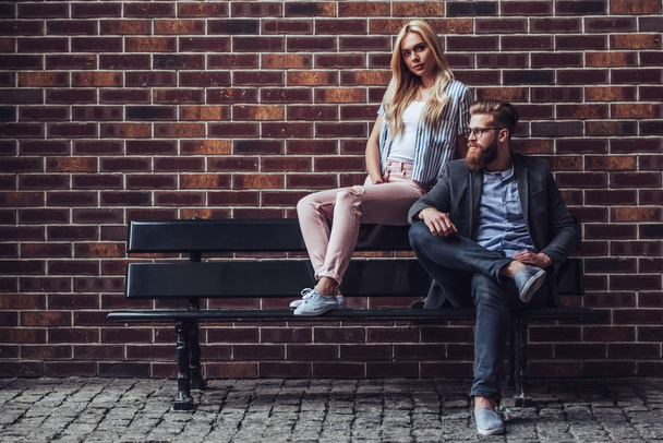 Stylish young couple is spending time together outdoors. Attractive woman and handsome bearded man are sitting on a bench on brick background in the city. - Foto, immagini