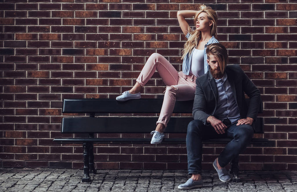 Stylish young couple is spending time together outdoors. Attractive woman and handsome bearded man are sitting on a bench on brick background in the city. - Фото, зображення
