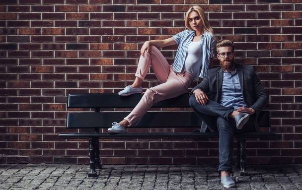 Stylish young couple is spending time together outdoors. Attractive woman and handsome bearded man are sitting on a bench on brick background in the city. - Fotó, kép