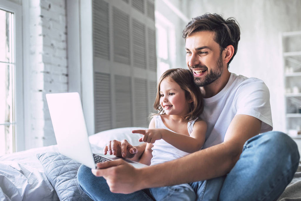 I love you, dad! Handsome young man at home with his little cute girl are lying in bed with laptop. Happy Father's Day! - Fotó, kép