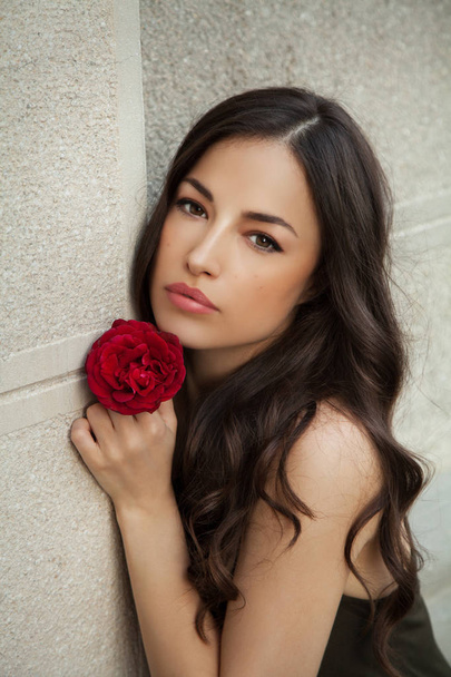 красива молода жінка тримає троянду близько до її обличчя, нахилена на білій стіні, зовнішній портрет
 - Фото, зображення