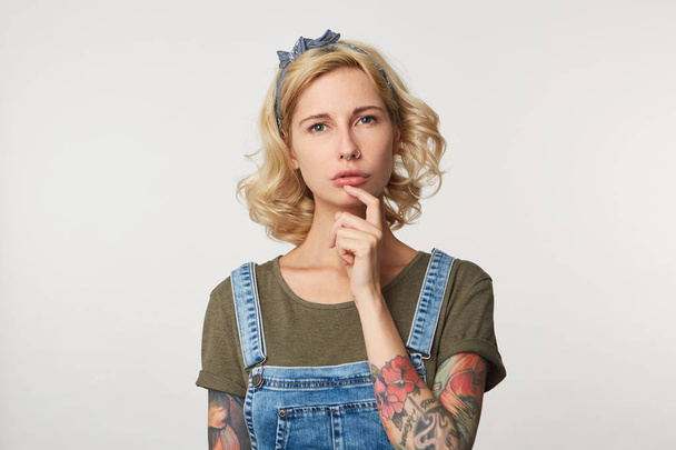 Realy? Studio shot of cute blonde female keep her finger under chin, weras denim overalls, thinking about her problems - Photo, Image