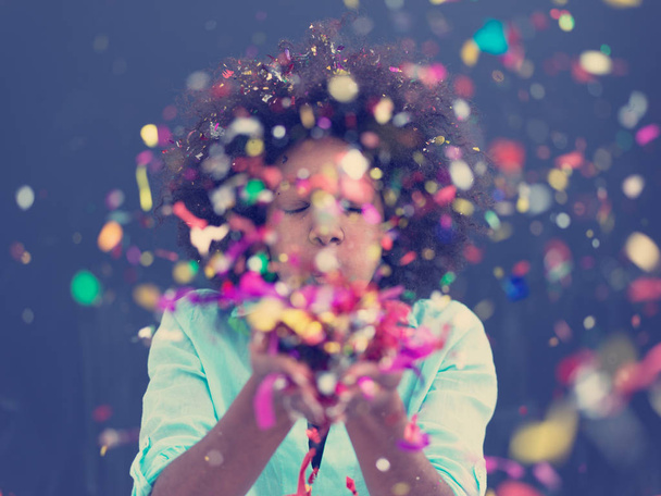 beautiful young black woman celebrating new year and Christmas party while blowing confetti decorations to camera isolated over gray background - Foto, Imagen