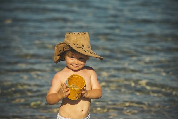 Mooie jongen met zee zand in een emmer in cowboy hoed tegen de blauwe zee. Zomer slang, warme sfeer. Gelukkig kind met een glimlach, een mooi gezicht, een close-up portret. Toerisme, vakantie met kind - Foto, afbeelding