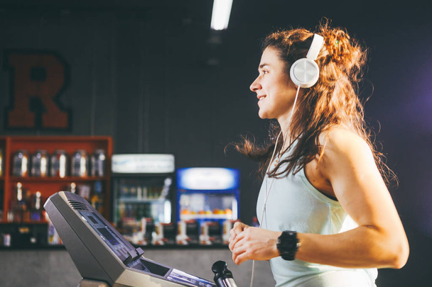 Theme is sport and music. A beautiful inflated woman runs in the gym on a treadmill. On her head are big white headphones, the girl listens to music during a cardio workout for weight loss - Photo, Image
