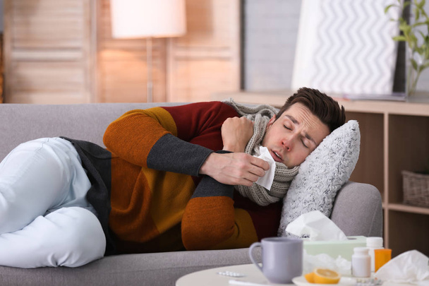 Sick young man with tissue suffering from cold on sofa at home - Foto, Bild
