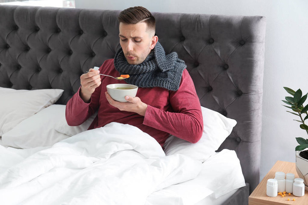 Sick young man eating broth to cure cold in bed at home - Foto, imagen