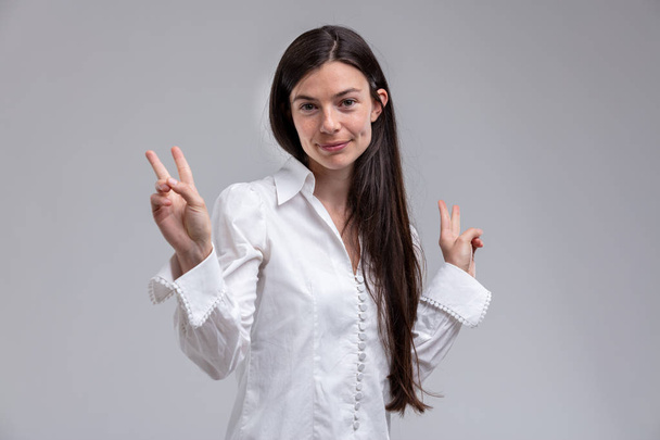 Portrait de jeune femme brune aux cheveux longs portant une chemise blanche montrant un signe de victoire
 - Photo, image