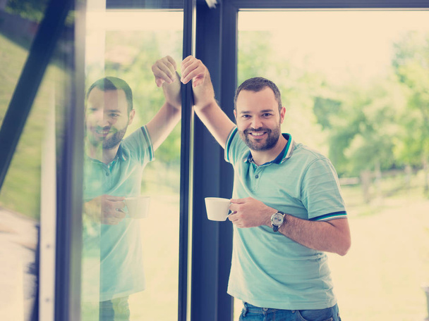 молодий красивий чоловік п'є ранкову каву біля вікна в своєму будинку
 - Фото, зображення